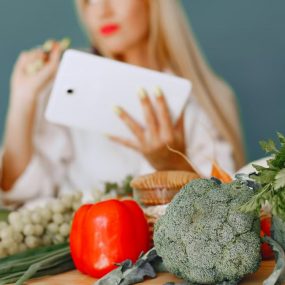 Blonde woman using tablet with fresh vegetables like broccoli and tomatoes in a kitchen setting.