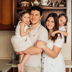 Satisfied man with daughter and woman with son embracing embracing each other while standing against wooden cupboard in kitchen and looking at camera
