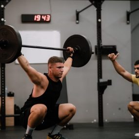 Muscular man lifting a barbell in a gym under his trainer's guidance, showcasing strength and focus.